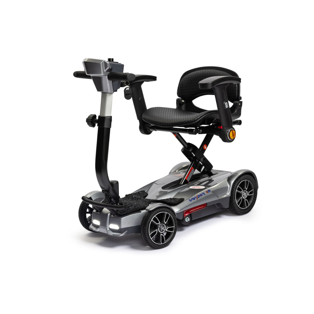 The EV Rider Transport 4M Travel Scooter is a compact, silver lightweight 4-wheel scooter with a black seat, armrests, and handlebar control panel, featuring a sturdy frame, shown against a white background.